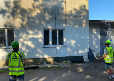 Dron podjetja SANDEX Fly & Clean čisti fasado stavbe, medtem ko ga nadzorujeta operaterja v zaščitnih jopičih.