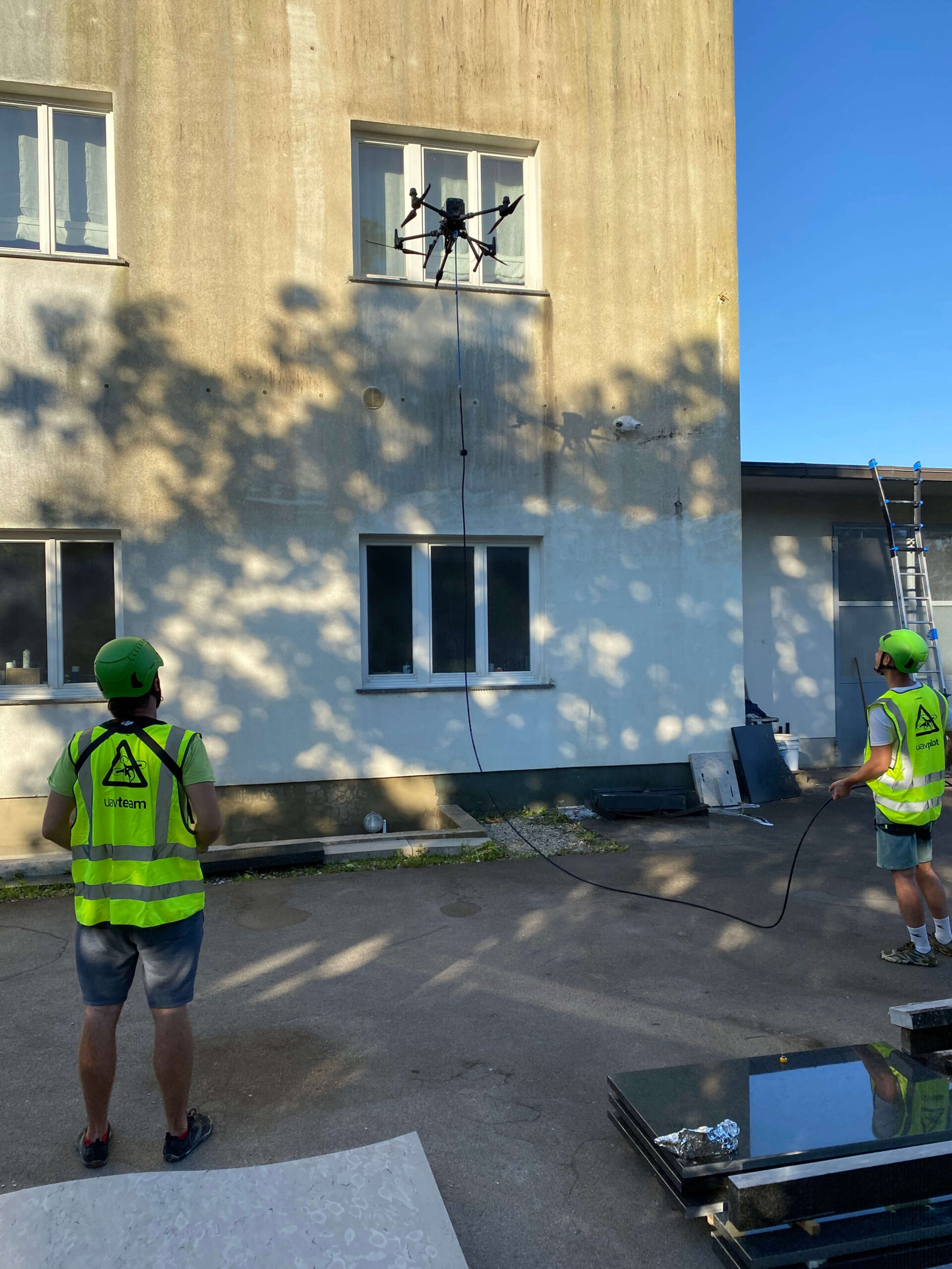 SANDEX dronsko čiščenje fasade v praksi Dron podjetja SANDEX Fly & Clean čisti fasado stavbe, medtem ko ga nadzorujeta operaterja v zaščitnih jopičih.
