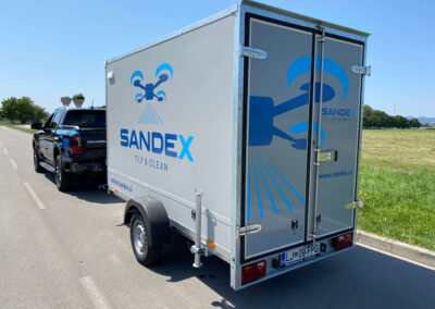 Prikolica podjetja SANDEX Fly & Clean s službenim vozilom za transport opreme za dronsko čiščenje objektov v Sloveniji.